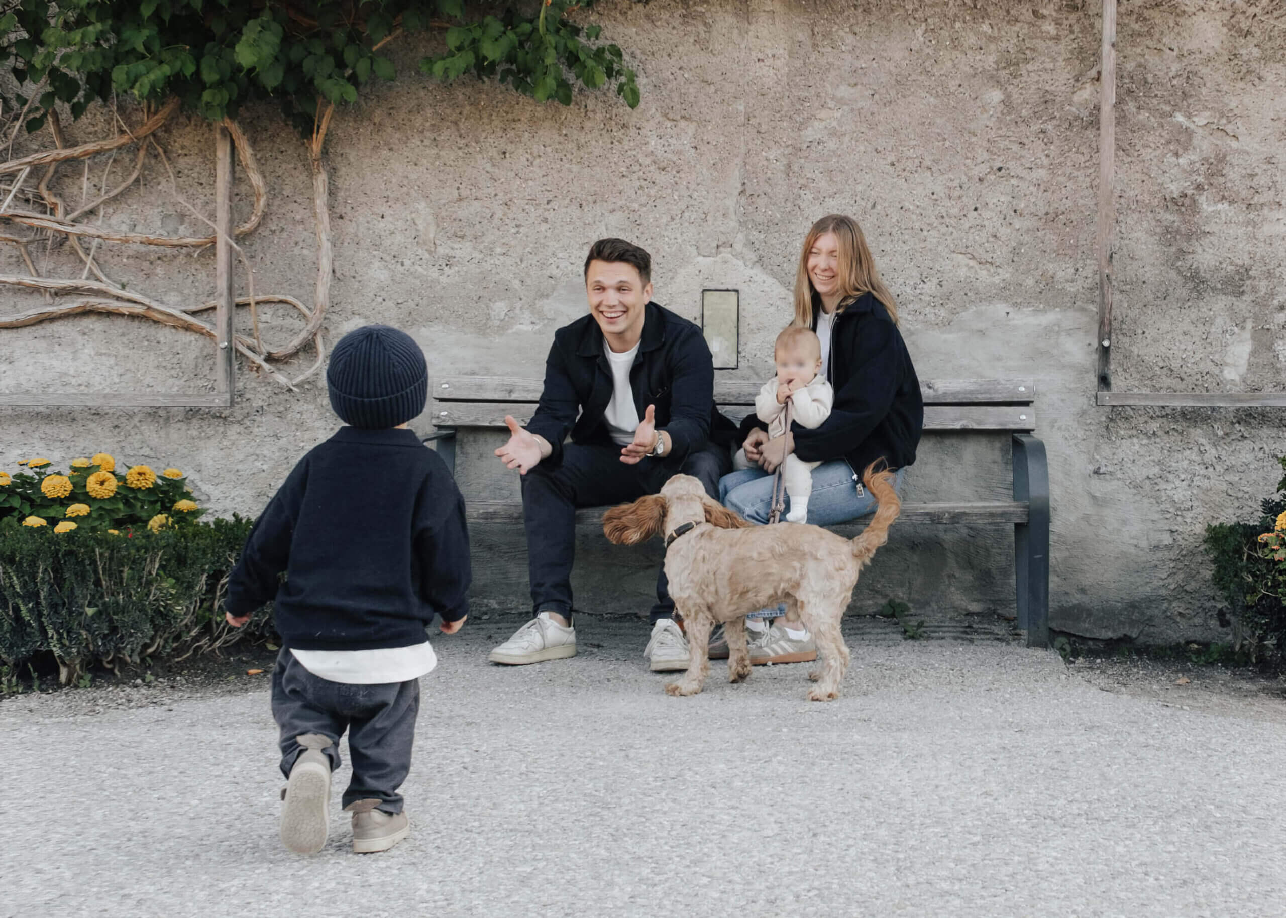 Familie mit Hund sitzt auf einer Bank, ein Kind läuft auf sie zu.