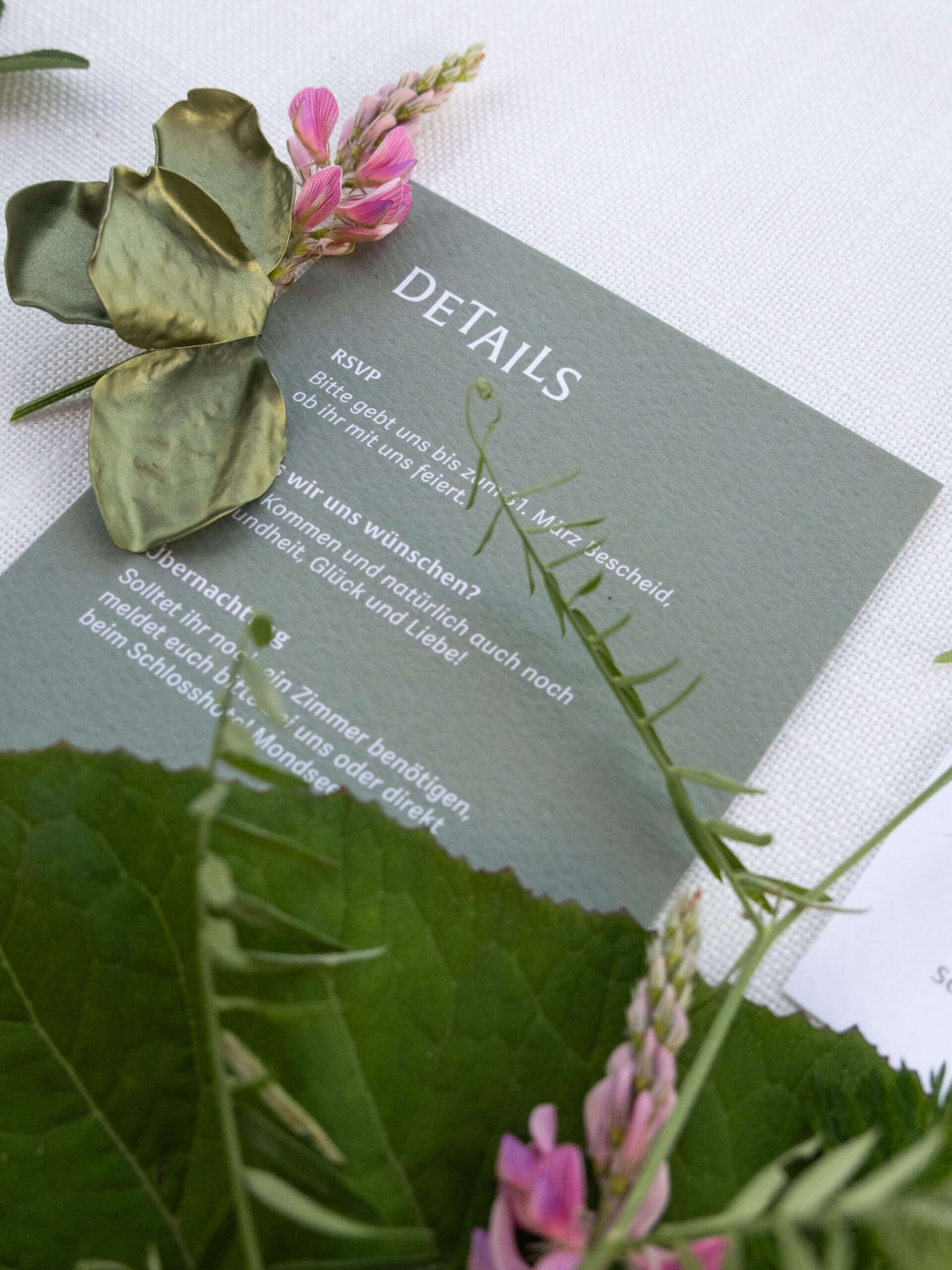 Hochzeitspapeterie mit "Details" und RSVP-Informationen, dekoriert mit grünen Blättern und rosa Blüten.
